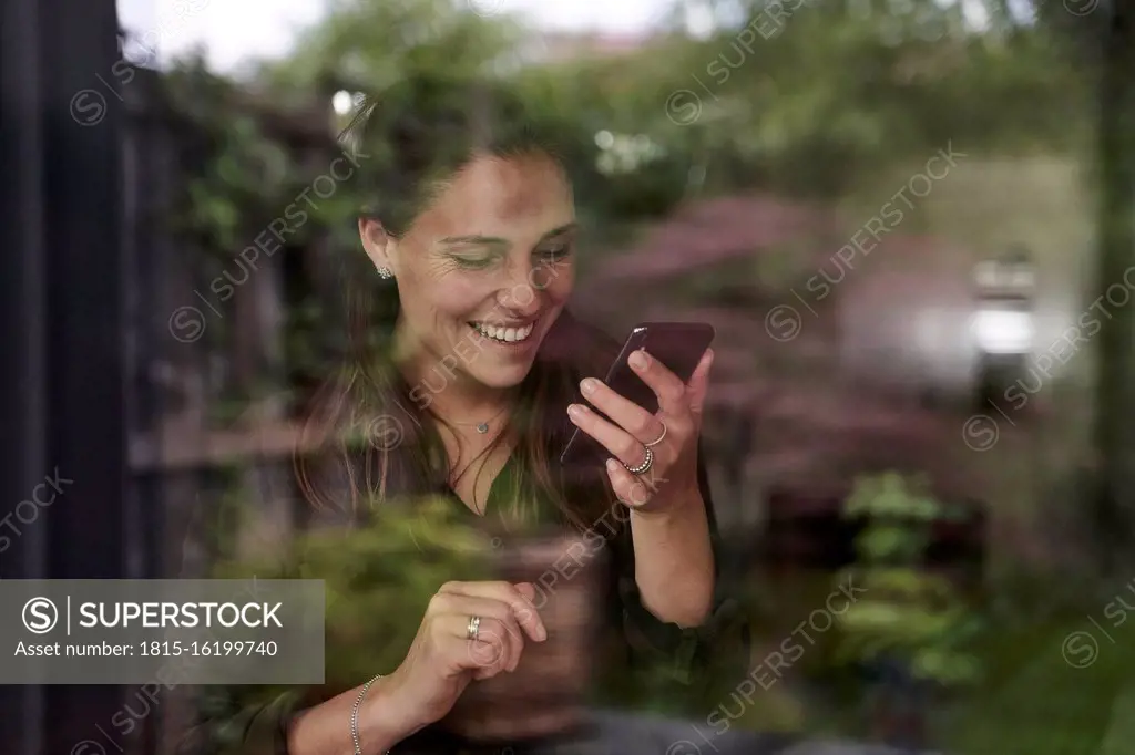 Smiling mid adult woman using mobile phone at home seen through window