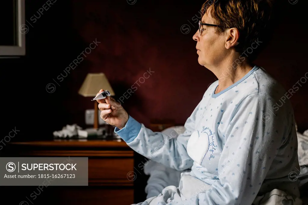 Ill senior woman looking at thermometer while sitting on bed at home