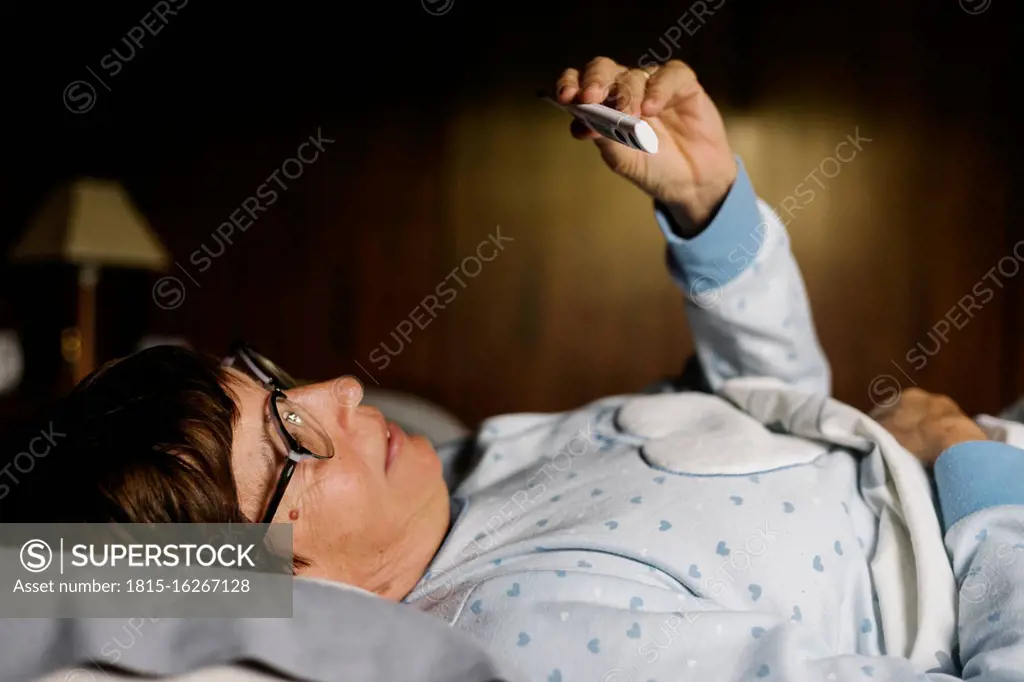 Elderly woman looking at thermometer while lying on bed at home