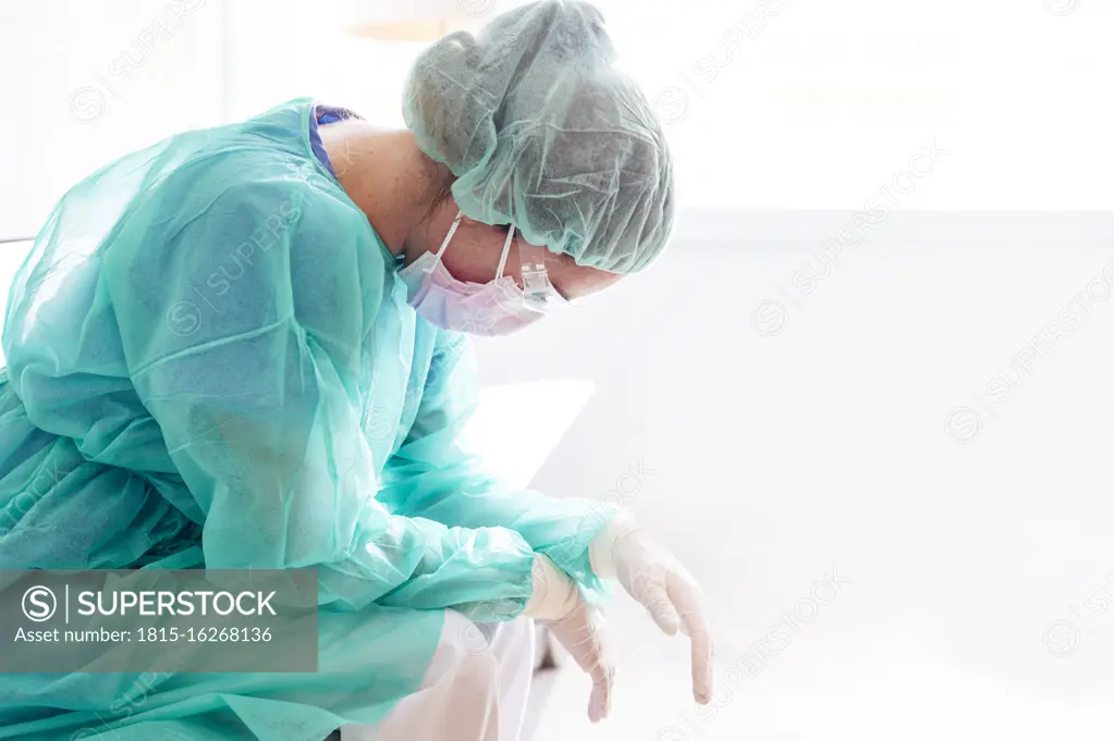 Sad female doctor wearing protective workwear sitting against window in hospital
