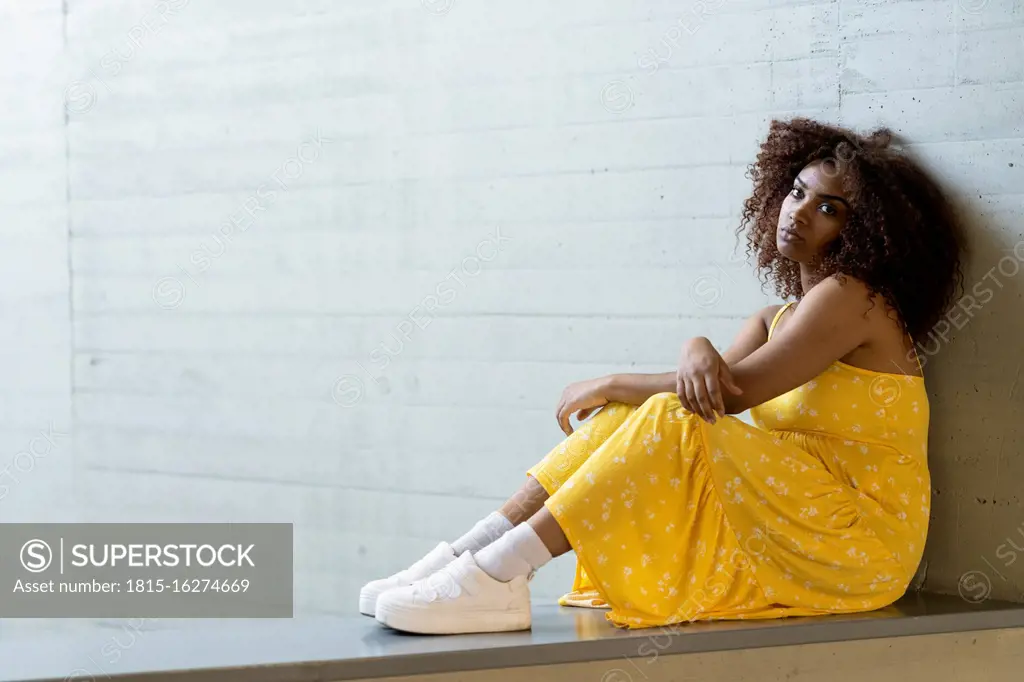 Sad woman with afro hair wearing yellow dress sitting on retaining wall