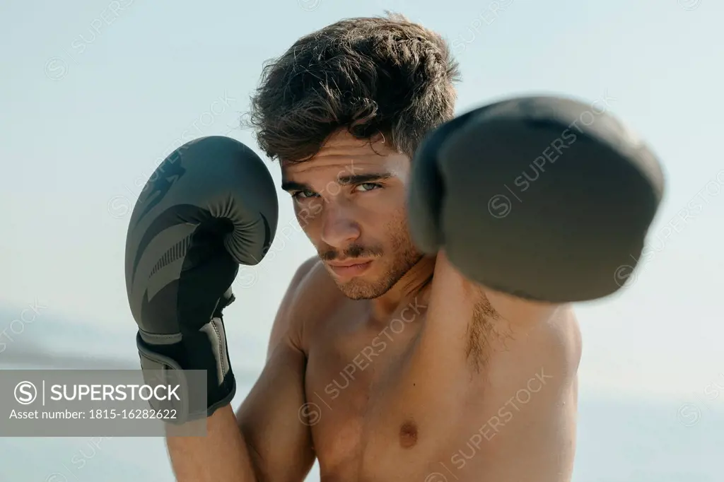 Close-up of shirtless boxer wearing gloves practicing boxing against clear sky