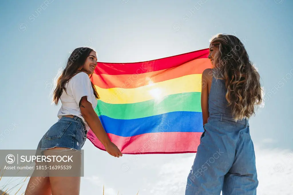 Two smiling young women holding an LGBT flag under blue sky