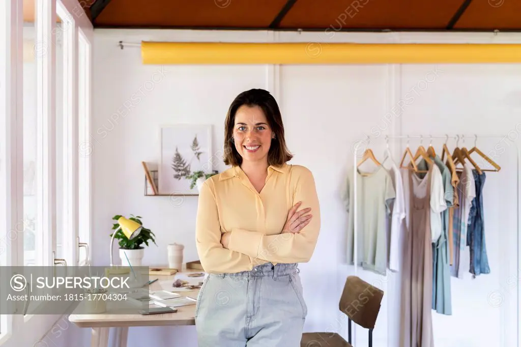 Smiling young female fashion designer with arms crossed leaning on table at clothing store