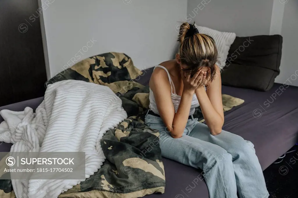 Despaired teenage girl sitting on bed, covering face with hands