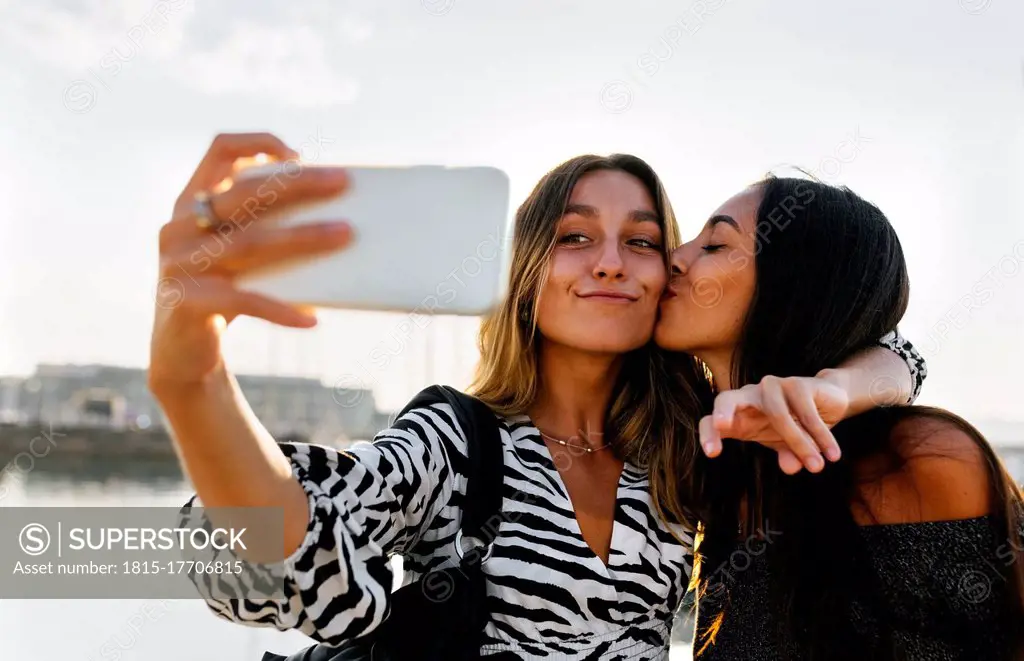 Young woman kissing friend while taking selfie against clear sky