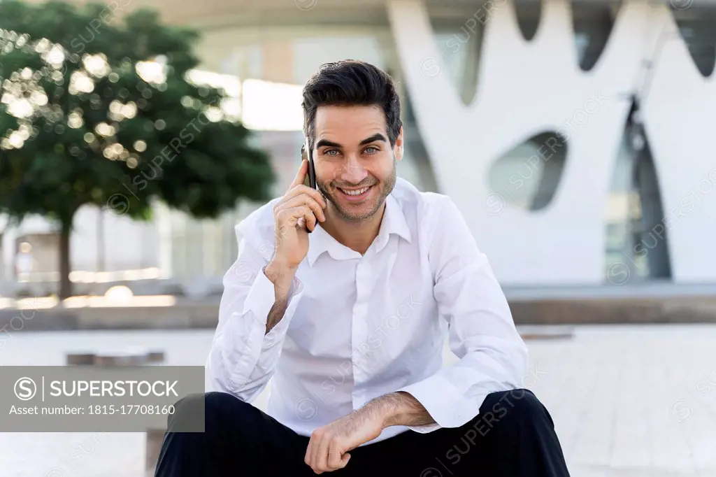 Smiling male entrepreneur talking over smart phone sitting against built structure in city