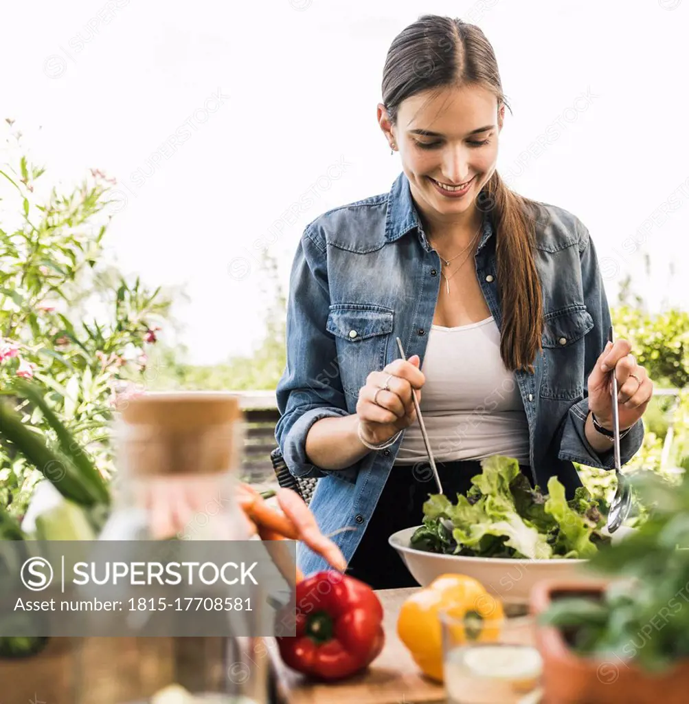 Smiling young woman making salad in bowl while sitting against clear sky at yard