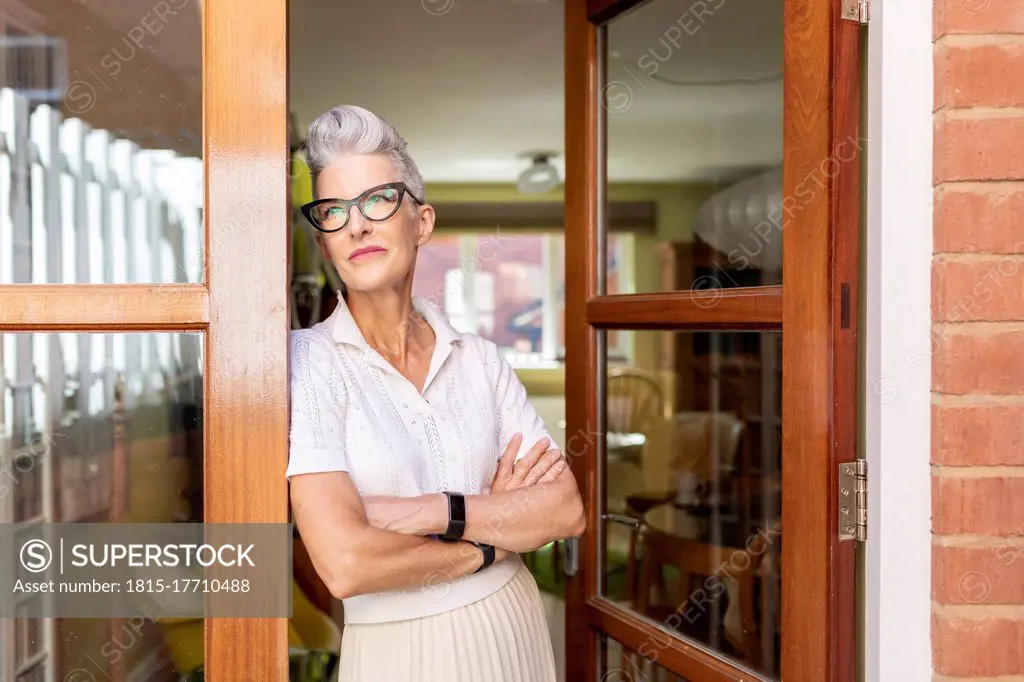 Contemplating senior woman with arms crossed standing on doorway at home