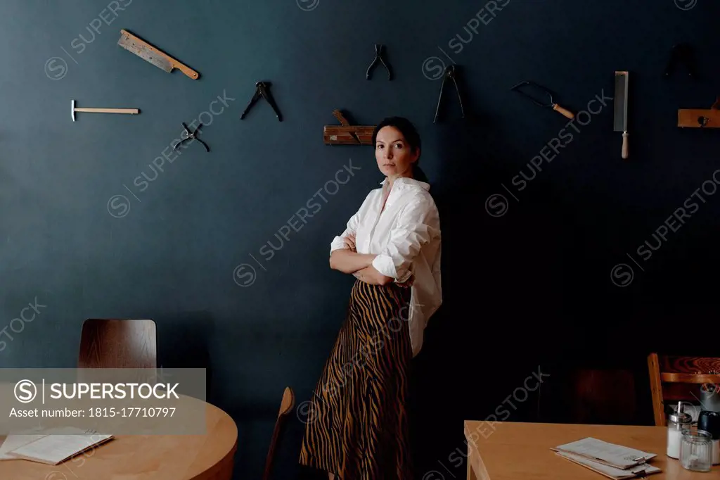 Female owner with arms crossed standing by work tool hanging on wall in coffee shop