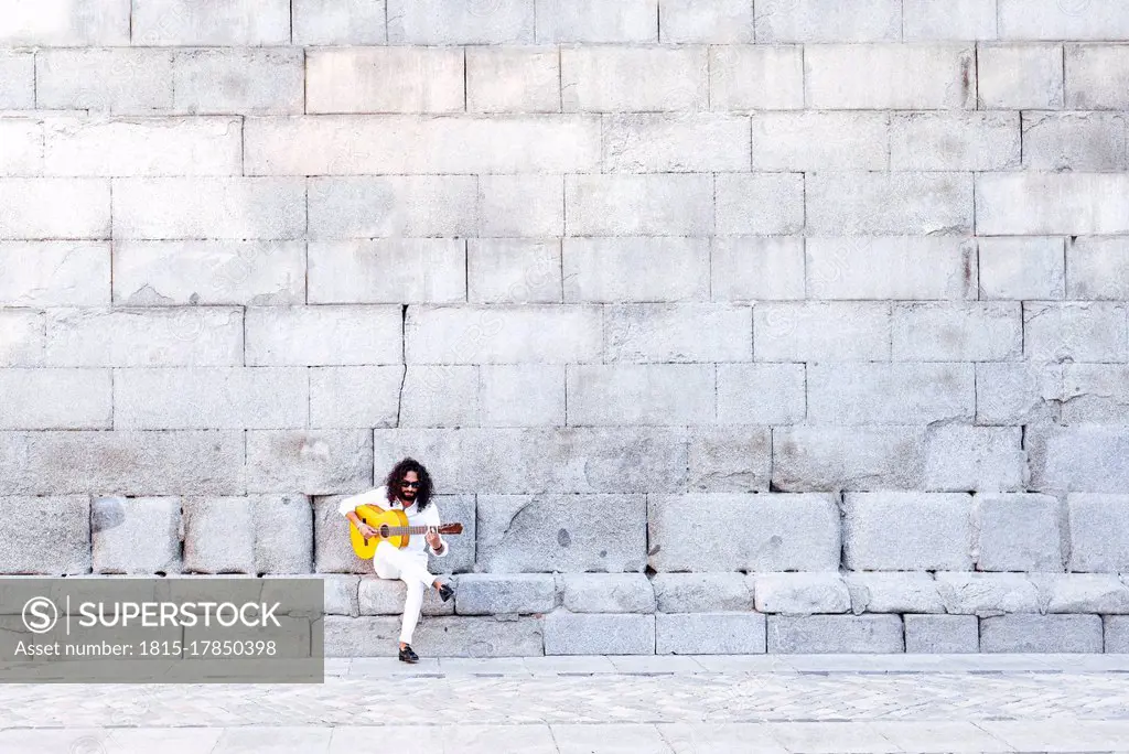 Flamenco guitarist playing guitar while sitting on bench against concrete wall