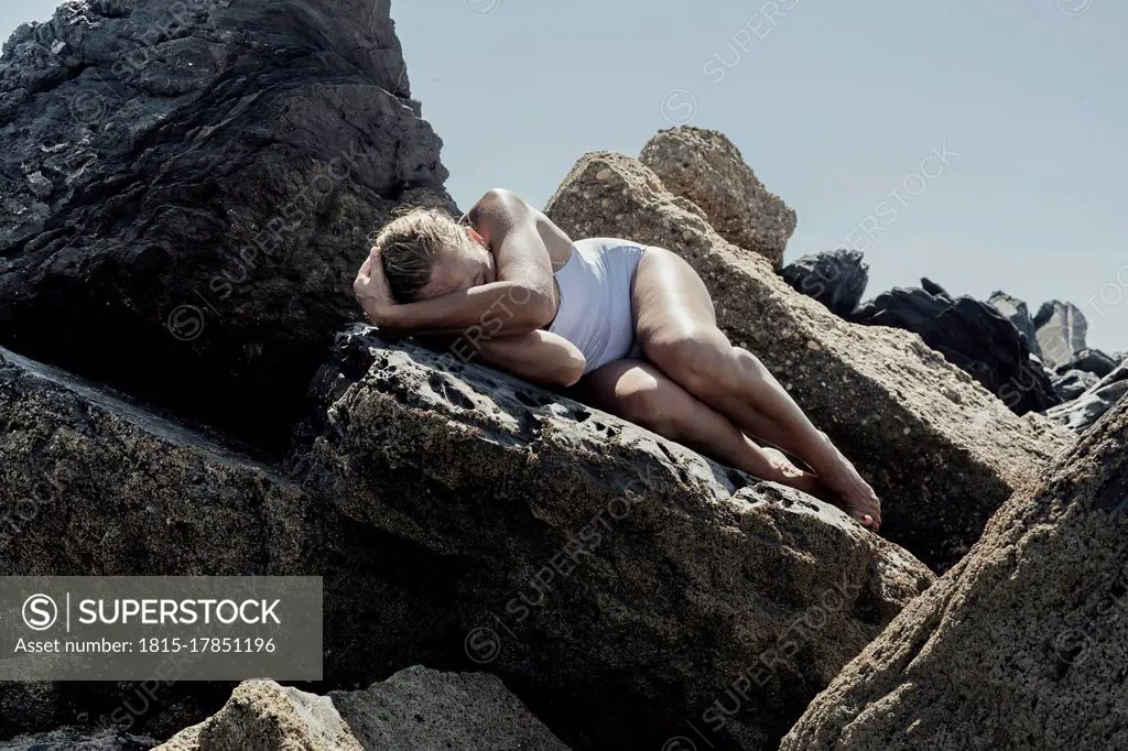 Senior woman wearing swimwear sleeping on rock at beach during sunny day