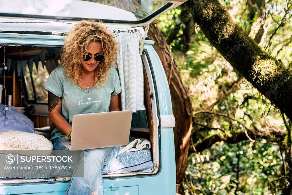 Woman wearing sunglasses using laptop while sitting on bed in vintage motor home