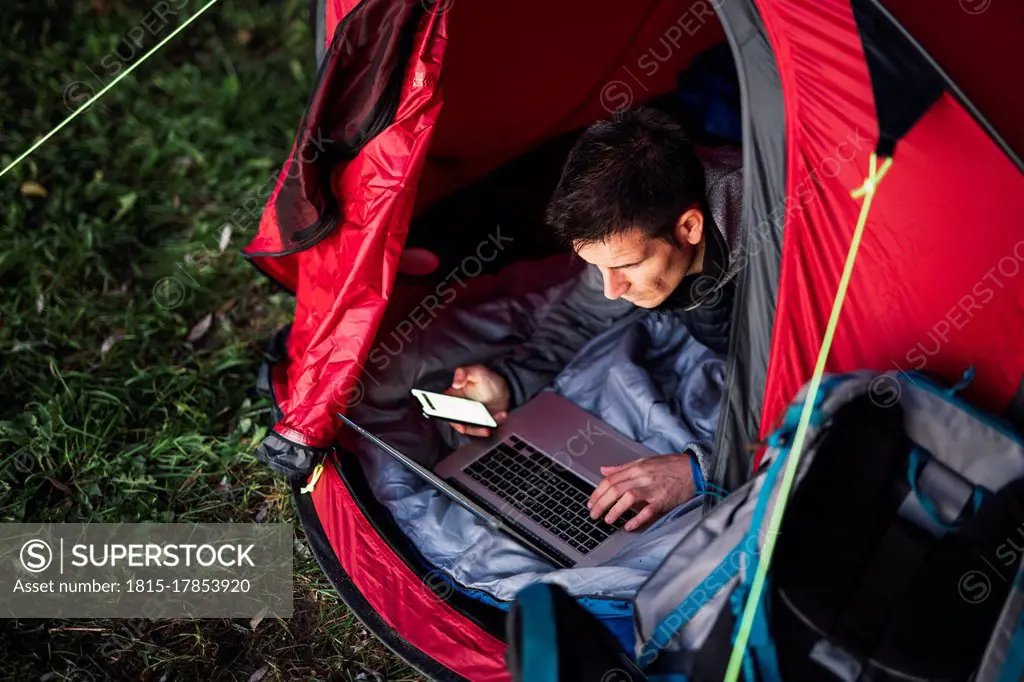 Man camping in Estonia, sitting in tent, using laptop