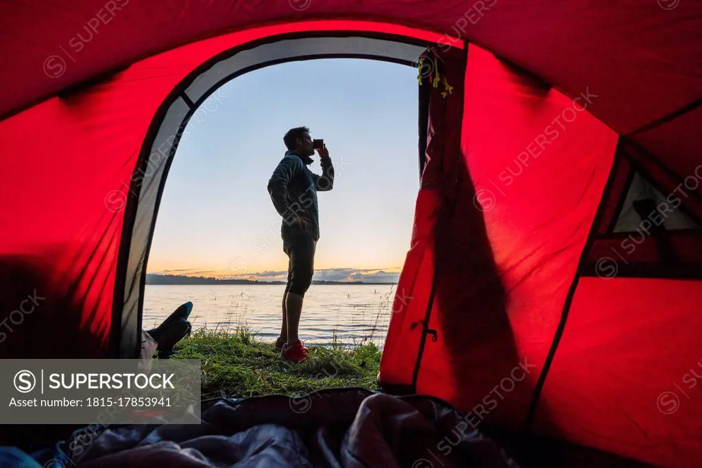 Man camping in Estonia, standing in front of tent, watching sunset