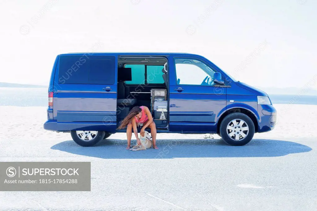 Woman petting dog while sitting on blue motor home at beach against clear sky