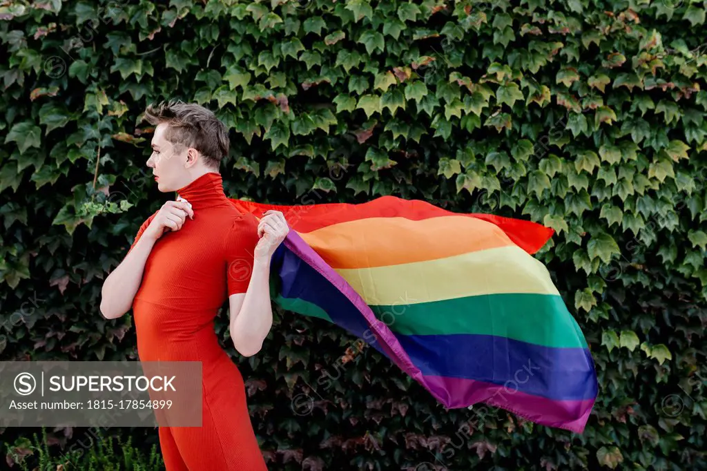 Non-binary person in red dress holding rainbow flag while standing against ivy wall