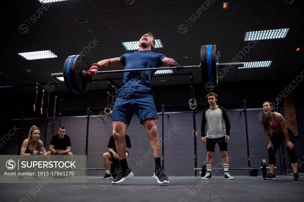 Adaptive athlete jumping while lifting barbell with people cheering in background at gym