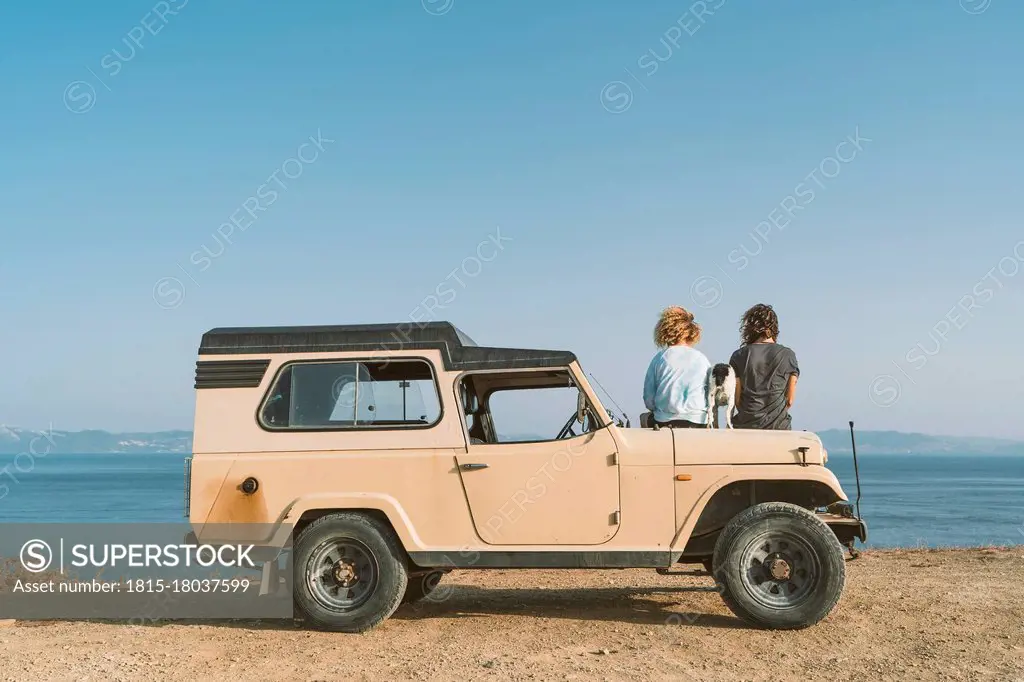 Young man and woman admiring sea view while sitting with dog on car at beach