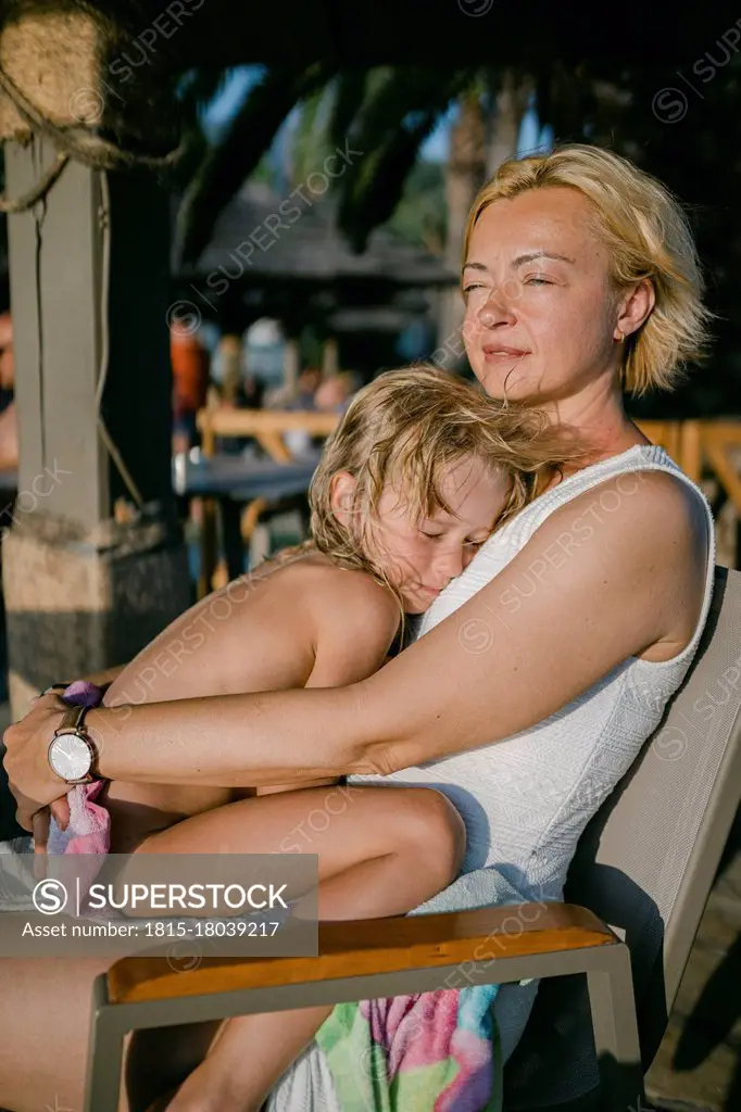 Daughter sleeping on smiling mother sitting on chair at hotel