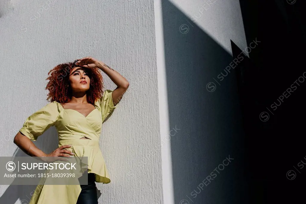 Fashionable woman shielding eyes while standing by day wall on sunny day