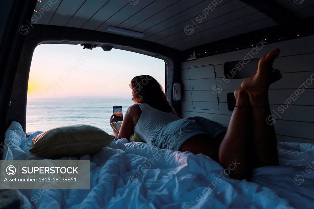 Woman taking photo of sunset through phone while lying in camper van at beach