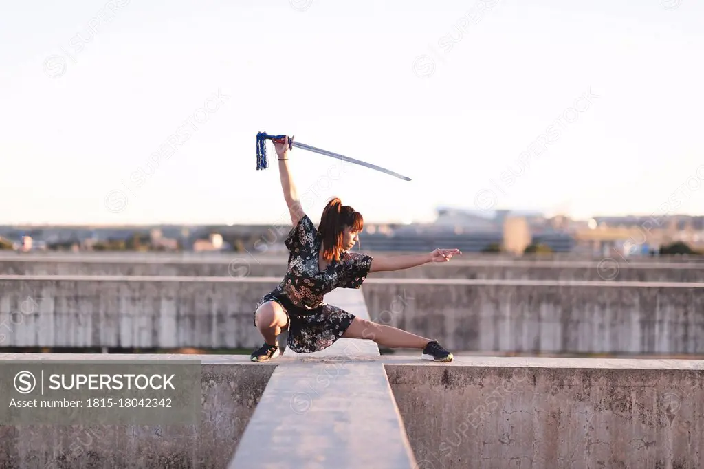 Young female athlete practicing sword on structure against clear sky during sunset