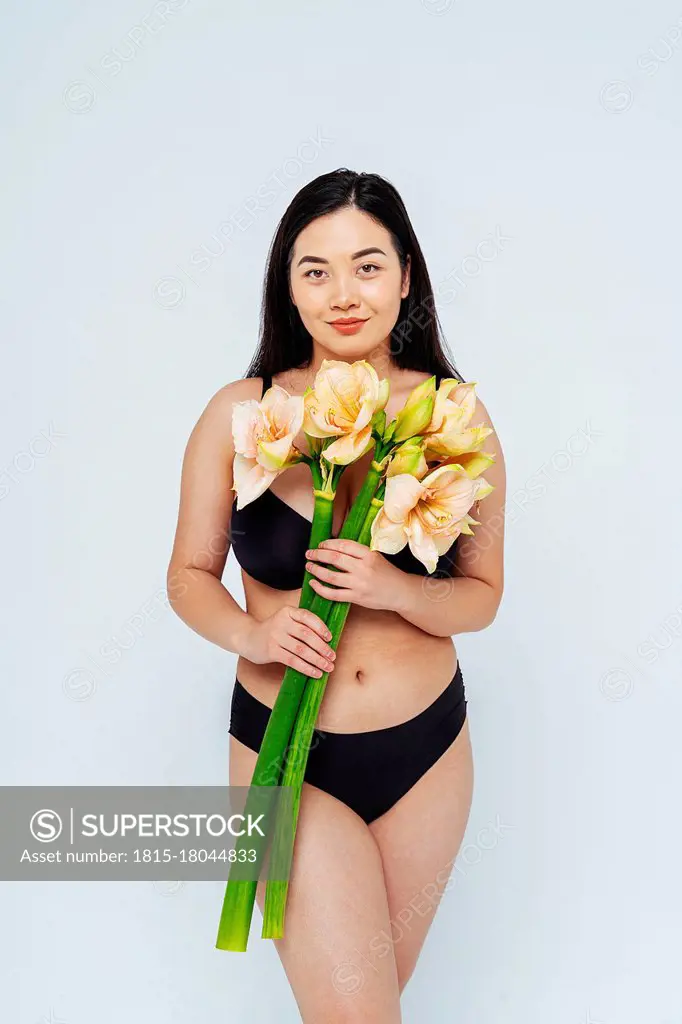 Young woman wearing lingerie holding bunch of flower while standing against white background