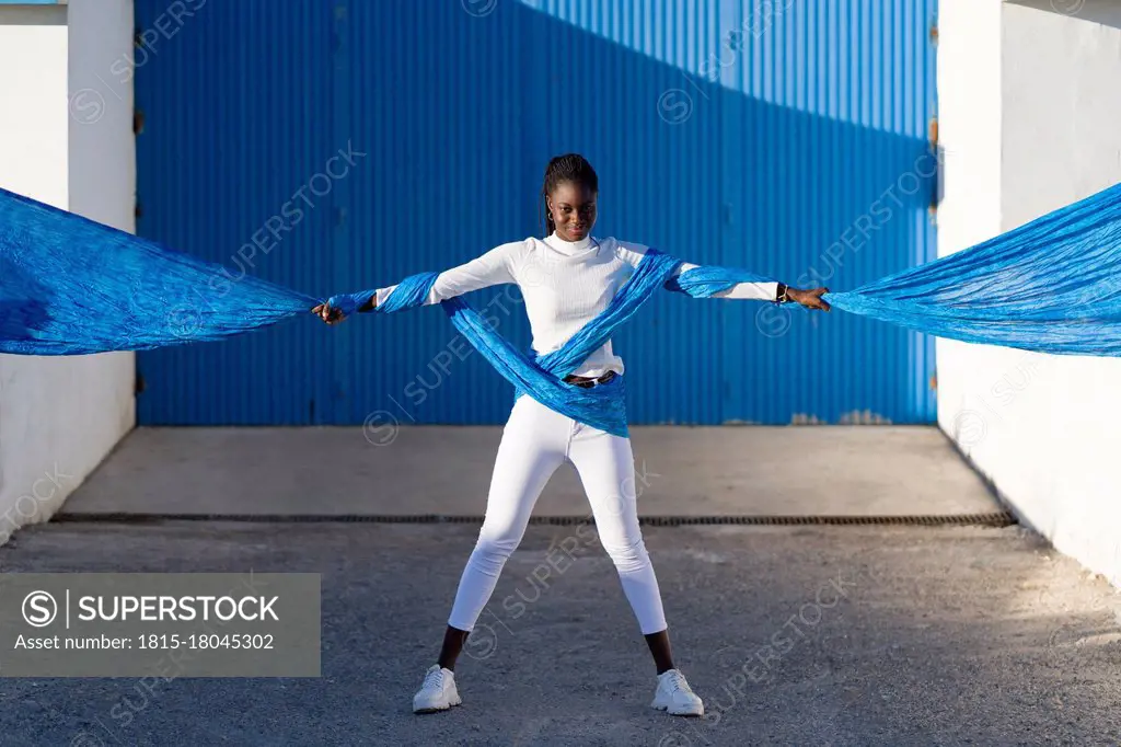 Teenage girl holding blue fabric while standing against corrugated wall on sunny day