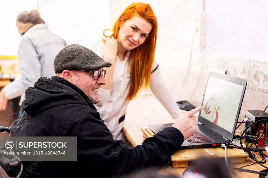 Smiling man using laptop by female caregiver in disabled center