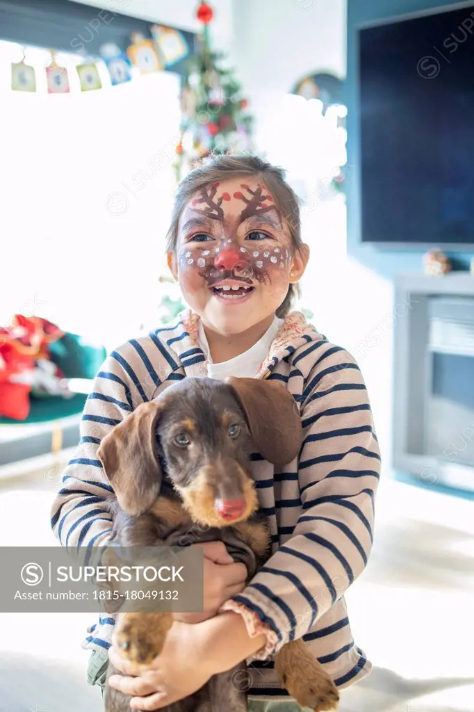 Smiling girl with painted face holding dog at home during Christmas