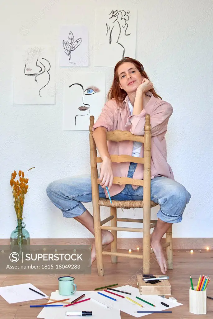 Smiling female artist with hand on chin at chair against wall in living room