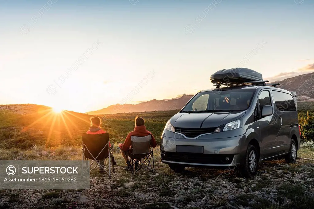 Man and woman sitting near camper van, looking at landscape at sunset
