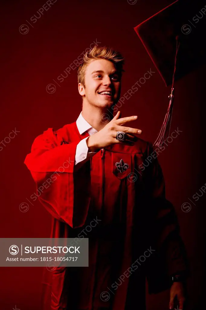 Male university student in graduation gown throwing mortarboard against red background