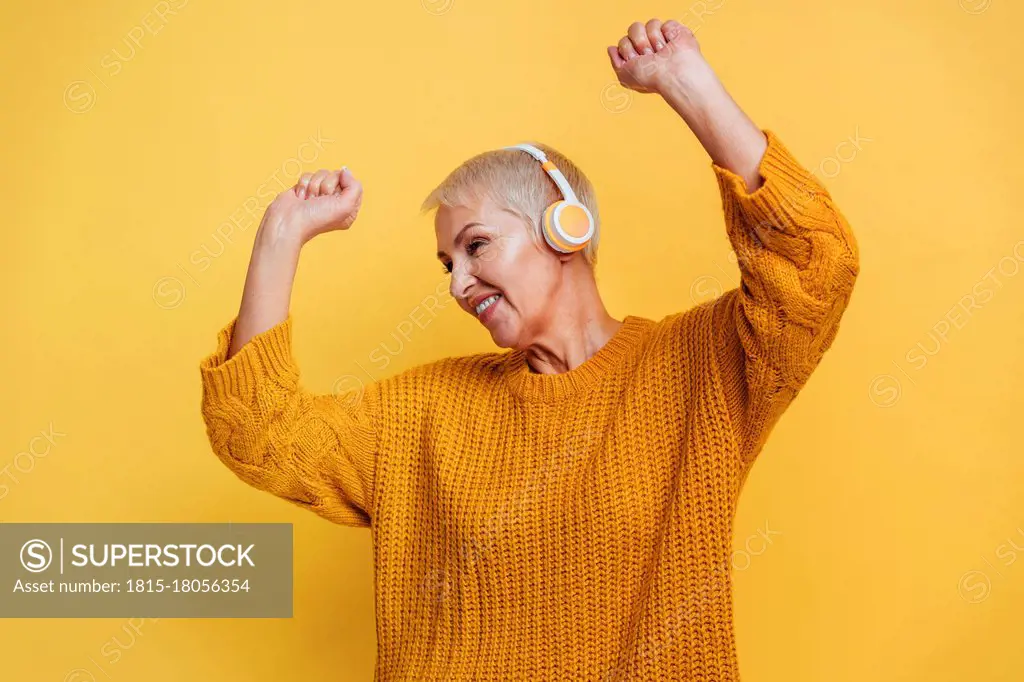 Carefree woman wearing headphones dancing with hand raised against yellow background