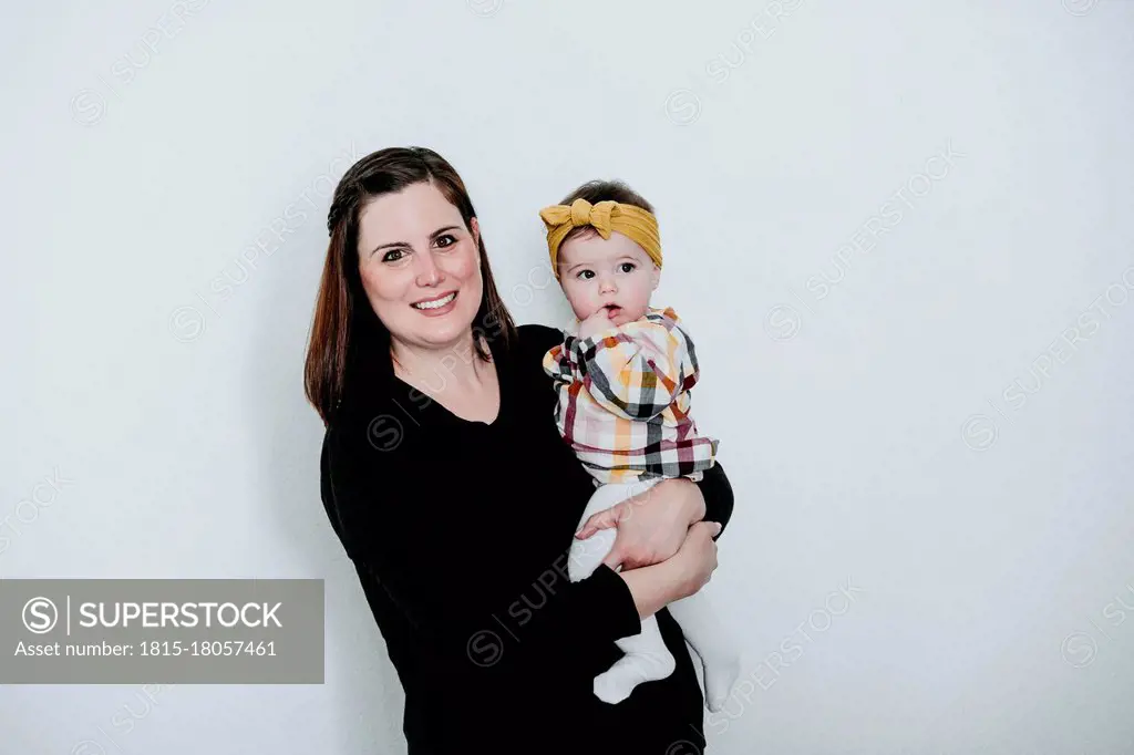Smiling mother carrying baby girl while standing against wall at home