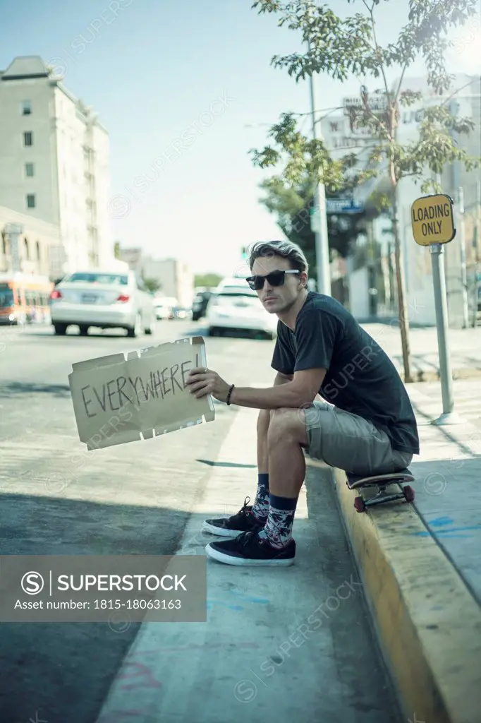 Young man holding signboard while sitting over skateboard on sidewalk