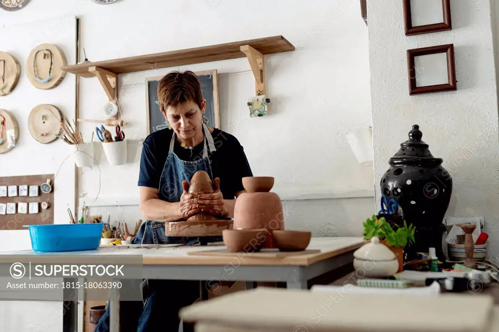 Mature female artist making pottery from brown clay in ceramic store