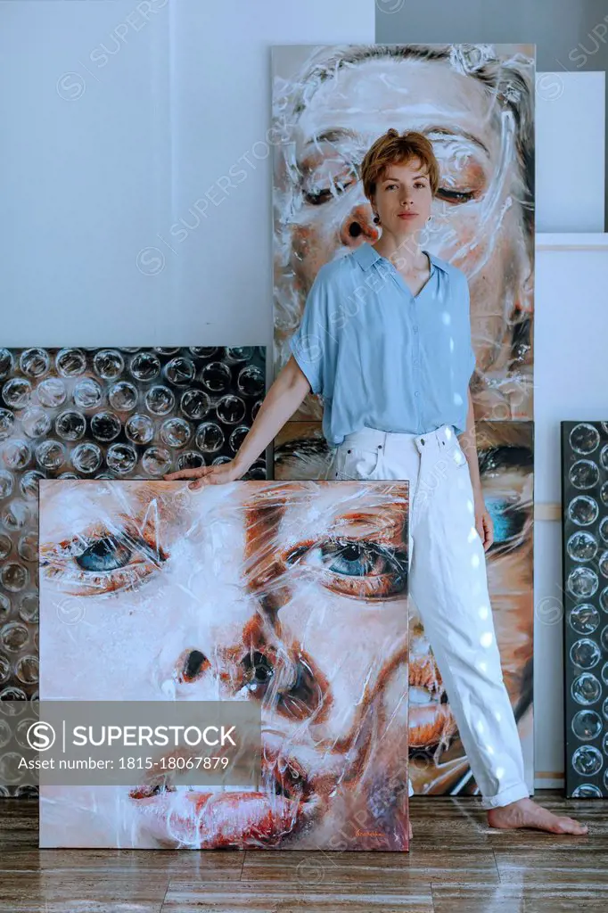 Confident mid adult female artist standing with various paintings in art studio