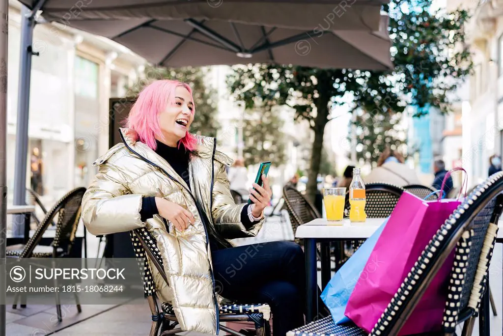 Pink hair woman using mobile phone while sitting with shopping bags at sidewalk cafe