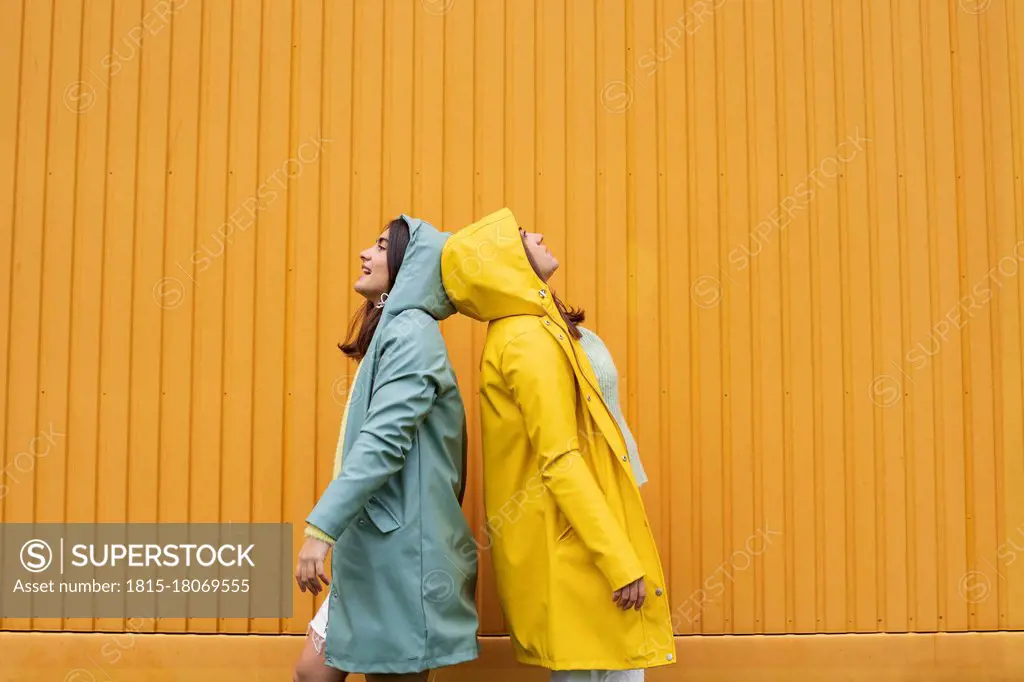 Young sister wearing blue and yellow raincoats standing back to back against wall