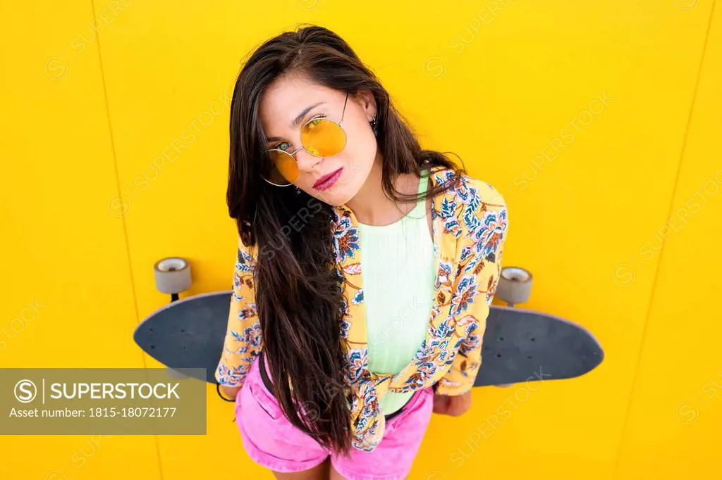 Portrait of young beautiful woman leaning on yellow wall with longboard in hands