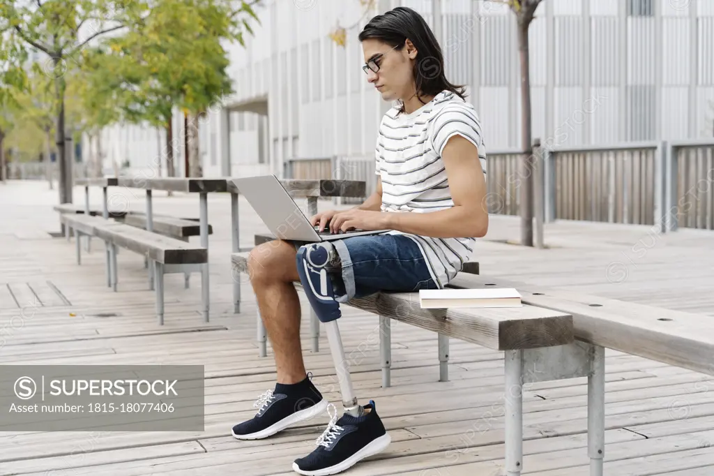 Contemplating man with artificial limb using laptop while sitting on bench in city