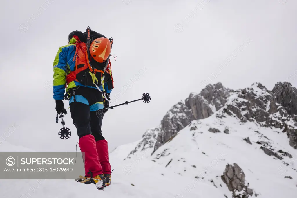 Mature man carrying backpack while walking on snowcapped mountain with crampon and hiking poles