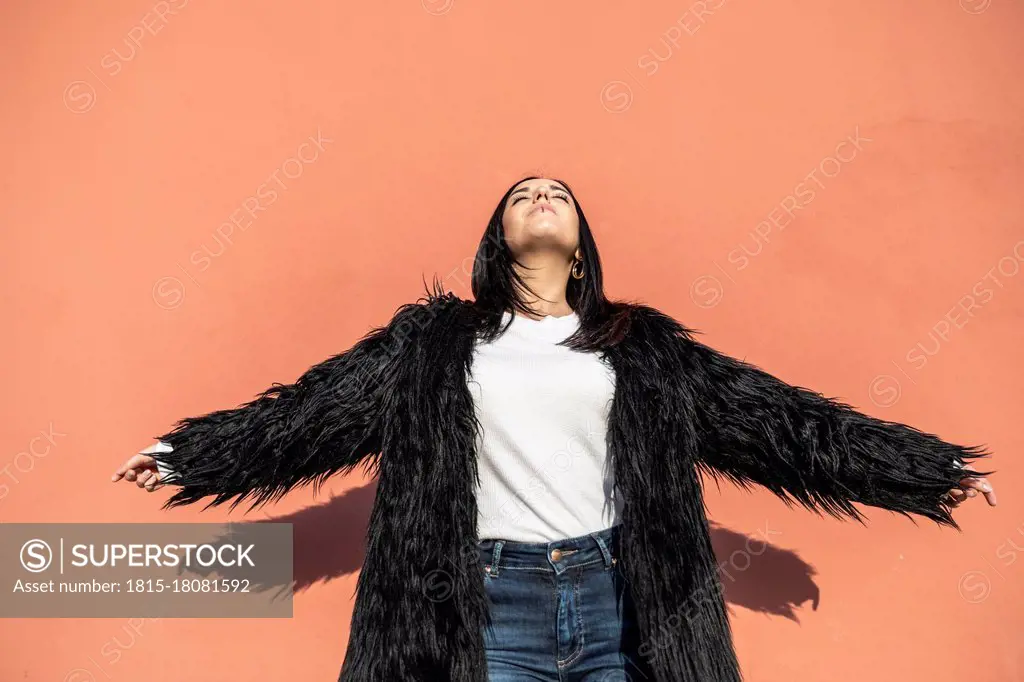 Carefree young woman with arms outstretched dancing against peach wall