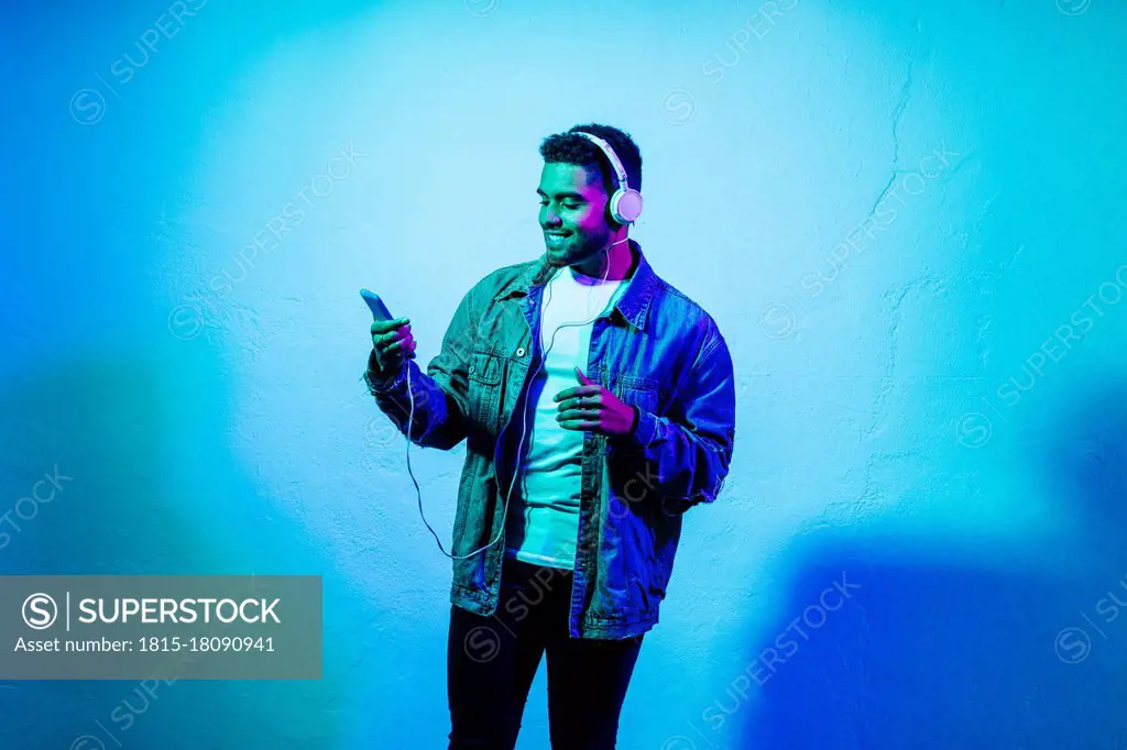 Smiling man with mobile phone listening music through headphones against colored background