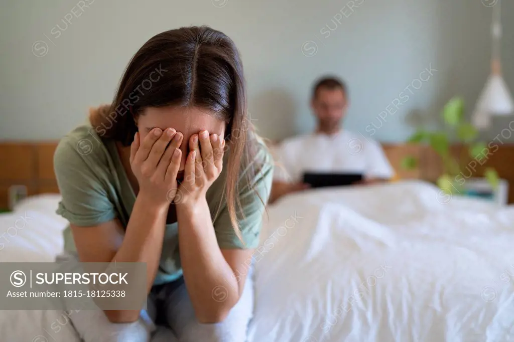 Desperate woman covering her face sitting on bed while boyfriend using digital tablet in background at home