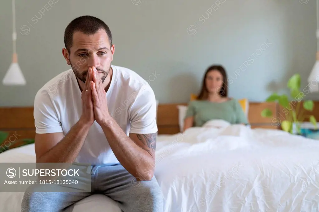 Serious man sitting on bed with girlfriend in background at home