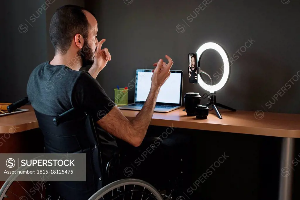 Vlogger filming through mobile phone with circular strobe tripod while sitting on wheelchair at studio