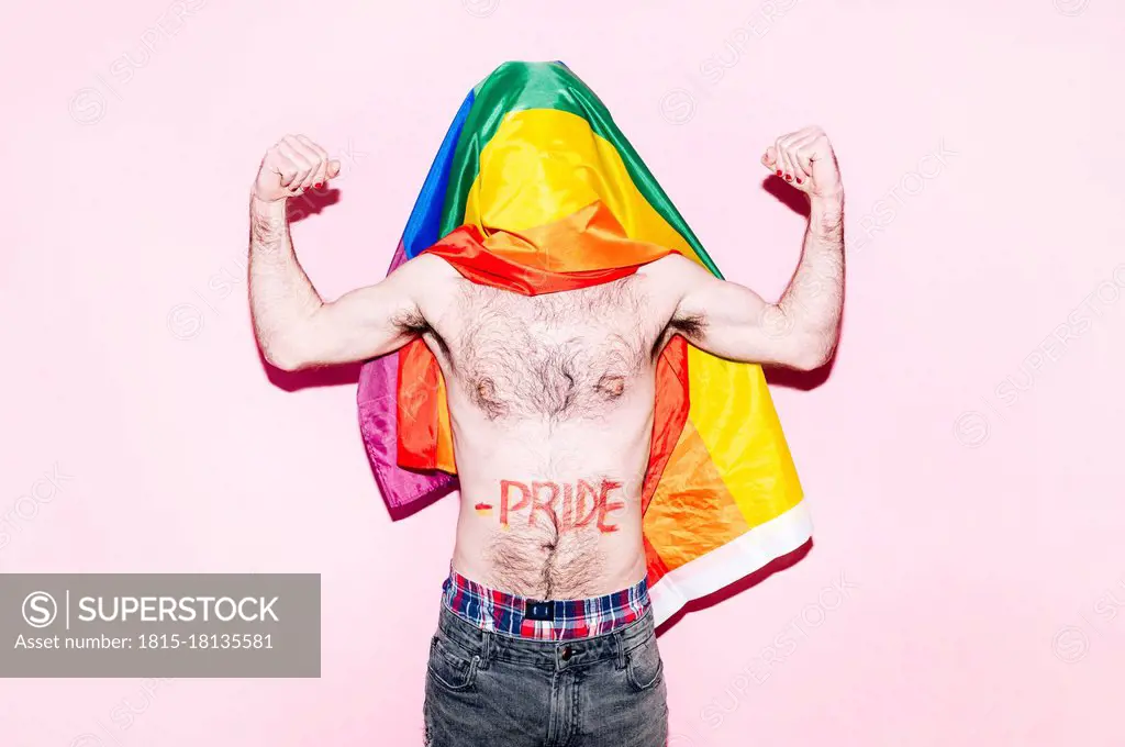 Covered face of gay man with rainbow flag flexing biceps standing against pink background
